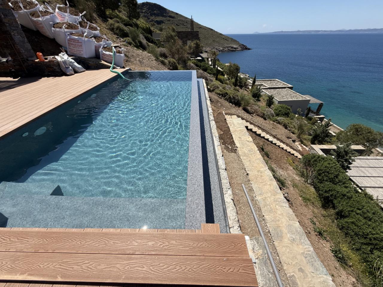 Hillside infinity pool with a wooden deck on the left, overlooking a blue sea and rugged coast; construction bags and equipment sit nearby against a rocky slope.