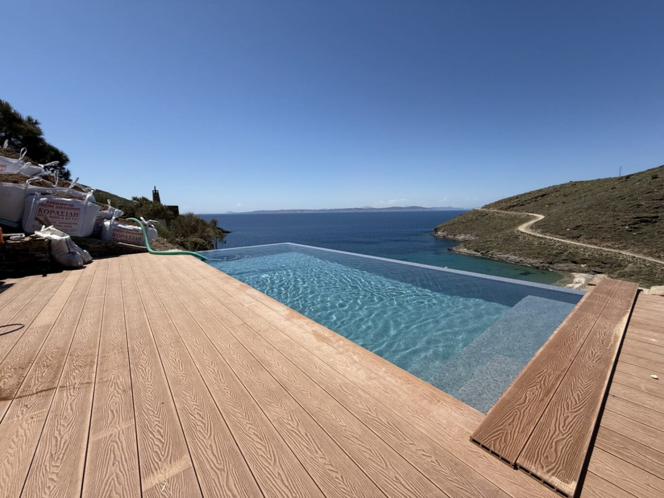 Infinity pool on a wooden deck overlooking the blue sea and distant islands, with a rocky hillside and road to the right.
