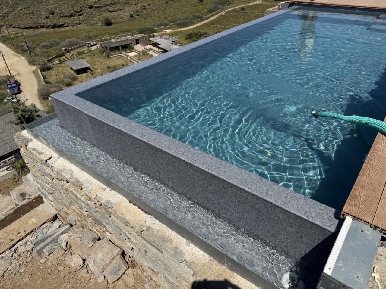 Rooftop infinity pool with blue water and stone coping overlooking a rural hillside village; construction debris nearby and a green hose in the pool.