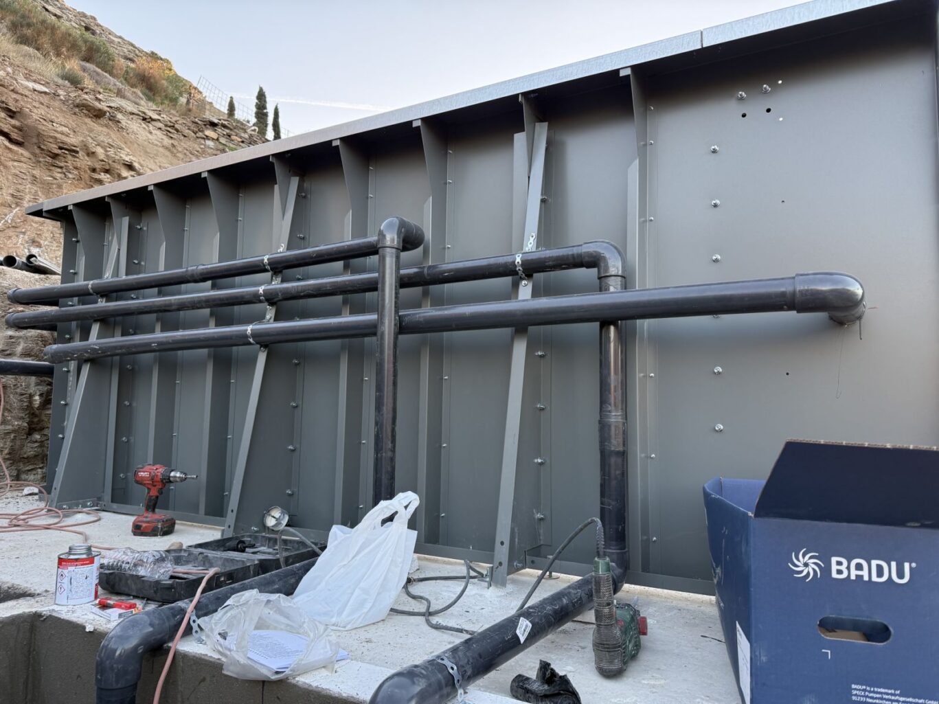 Grey industrial wall with large black pipes running horizontally; construction tools and debris on the ground nearby at a worksite.