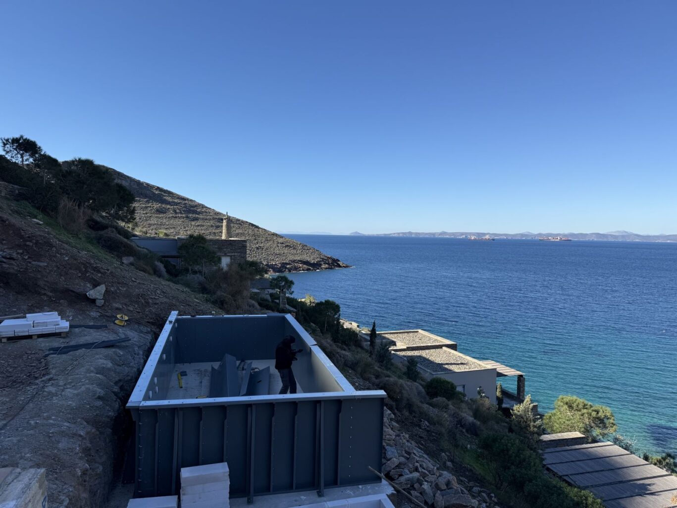 Construction area on a rocky hillside overlooking the blue sea, with a partially built metal-framed structure and a worker inside.