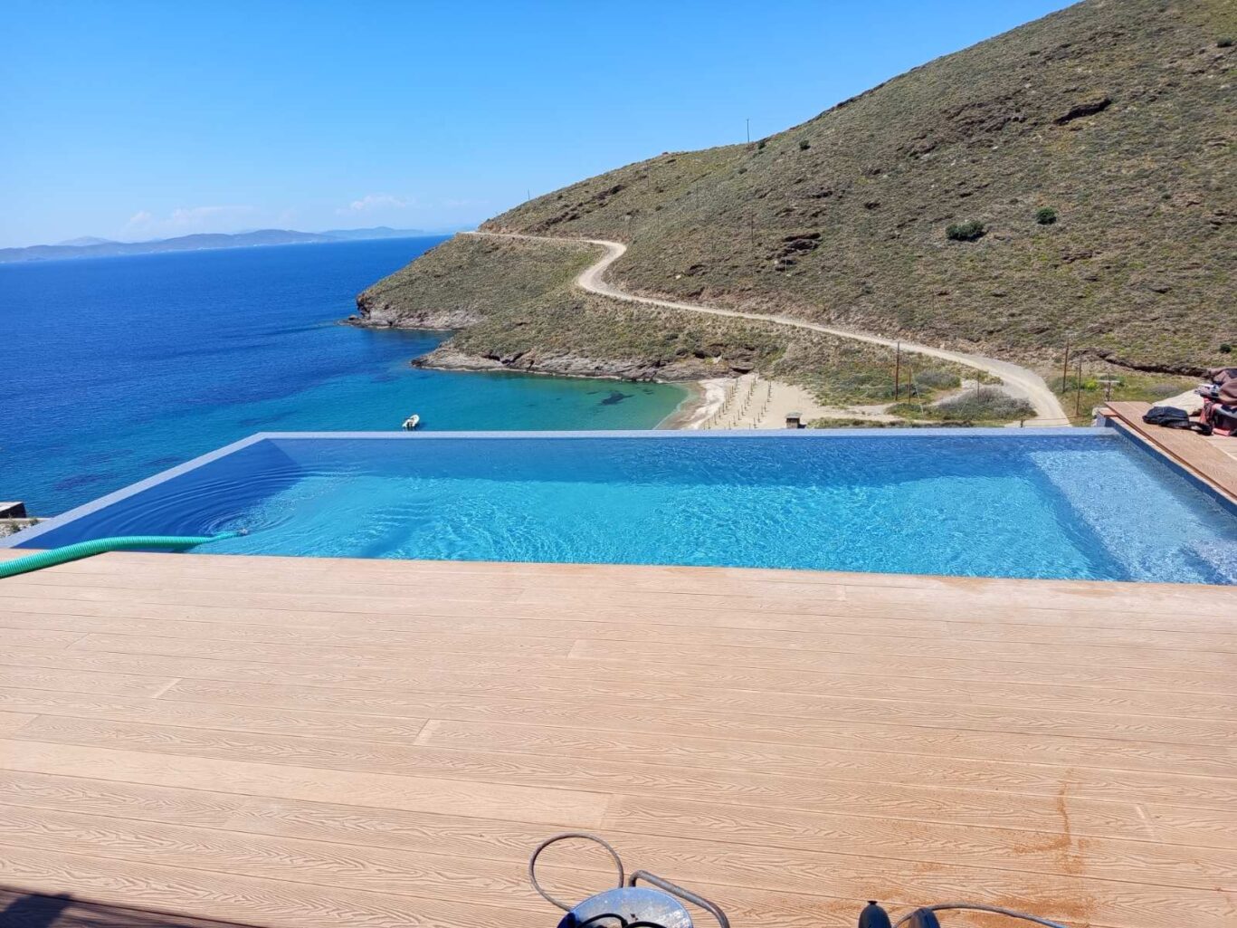 Infinity pool on a wooden deck overlooking the blue sea and a winding hillside road in the distance.