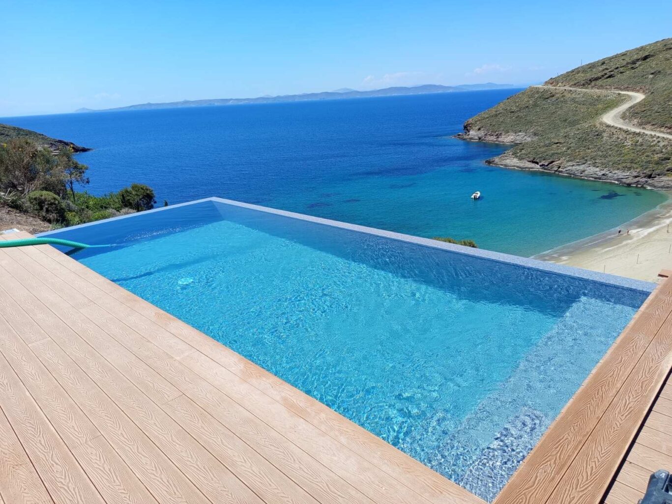 Infinity pool on a wooden deck overlooking a blue sea and coastline with a beach and hills in the distance.