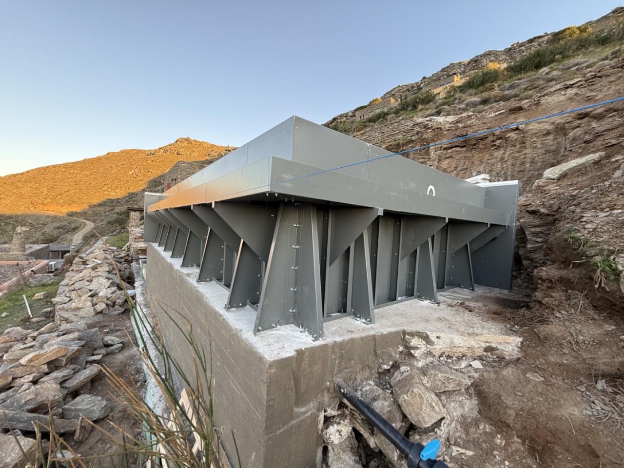 Steel-framed structure on a concrete foundation at a rocky hillside construction site, with diagonal braces supporting a long metallic beam.